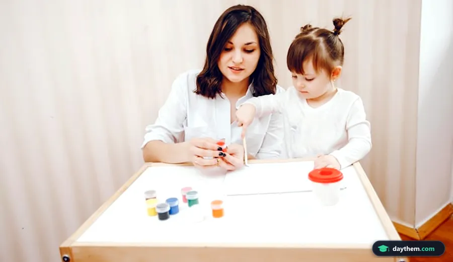 Bé gái và một phụ nữ trẻ ngồi cùng nhau bên bàn, đang vẽ tranh trên giấy bằng màu vẽ nhiều màu sắc.