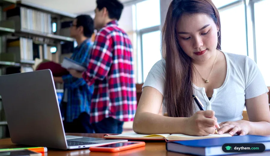 Hai học sinh nữ ngồi cạnh nhau trong thư viện, sử dụng máy tính xách tay và sách vở để học tập và tra cứu thông tin tuyển sinh đại học