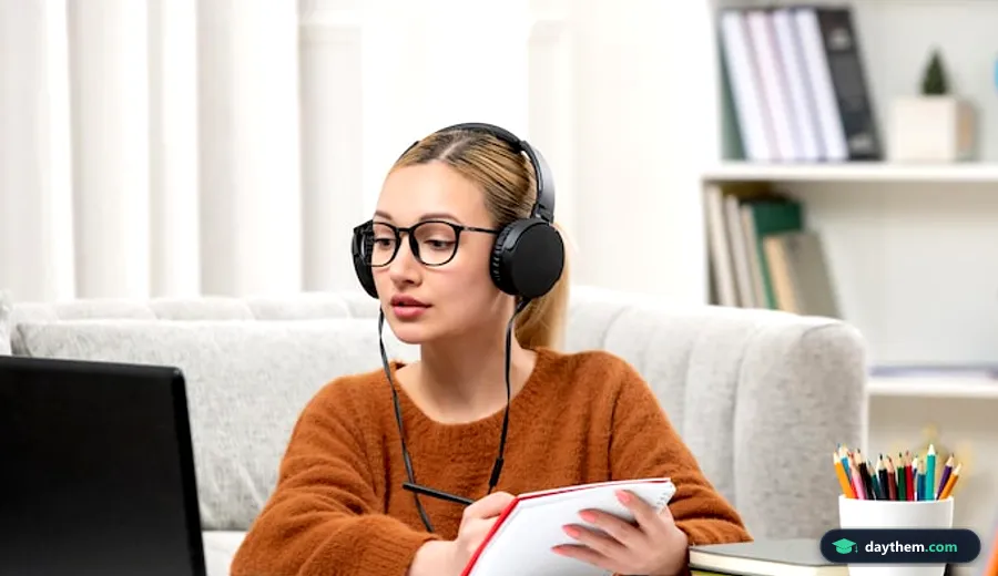 Học viên nữ đeo kính và mặc áo len đang tự học tiếng Anh trực tuyến trên máy tính và ghi chép vào sổ tay